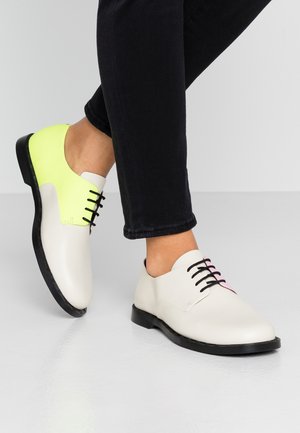 Persoon draagt witte leren veterlaarzen met zwarte zolen, waarbij één schoen een neongele hiel heeft, en zwarte broek die boven de enkels is afgeknipt.