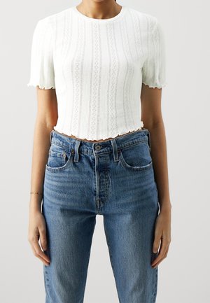 Personne portant un crop top en maille blanc à manches courtes avec des bords festonnés et un jean bleu taille haute, debout devant un fond uni.