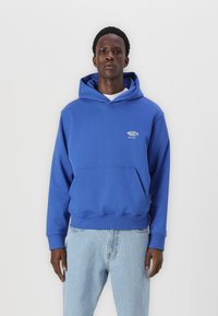 Blaues Baumwoll-Hoodie mit einer Vordertasche und Logodetails. Kombiniert mit hellblauen Jeans. Schlichtes Design und lockere Passform.