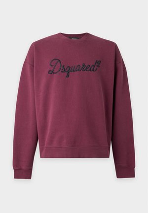 Νέος άνδρας φορώντας μωβ φούτερ Dsquared2, φθαρμένο μπλε τζιν και μαύρα αθλητικά παπούτσια, στέκεται μπροστά σε μια απλή φωτεινή επιφάνεια.