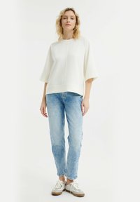 Top bianco con maniche raglan, abbinato a jeans in denim azzurro chiaro. Il modello indossa sneakers beige e bianche. Design semplice e pulito.