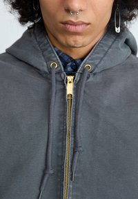 Grå hættetrøje med en guldlukning, snore og store huller. Underneden er en blå plaidskjorte delvist synlig, med mørke knapper.