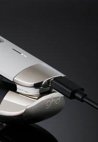 Gros plan sur un lisseur ghd argent et blanc avec un cordon d'alimentation noir branché, sur un fond sombre.