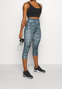 Leggings a vita alta, con un motivo a leopardato in turchese e grigio, realizzati in materiale elasticizzato, abbinati a scarpe da atletica nere e una borraccia.