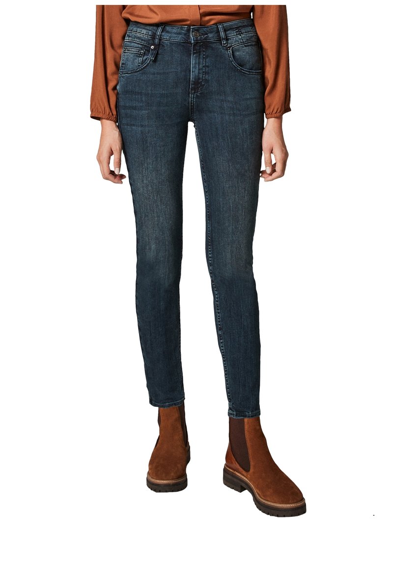 Personne portant un jean slim bleu foncé, un chemisier couleur rouille et des bottines en daim marron, debout sur un fond blanc.