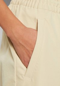 Beige fabric shorts with an elastic waistband and side pocket. The texture is smooth, with visible stitching along the pocket opening.