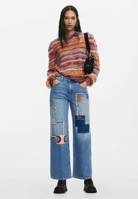 Maglione a righe multicolore in lavorazione a maglia con maniche lunghe abbinato a jeans a gamba larga blu con vari patch di tessuto e dettagli ricamati.