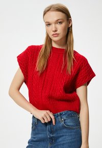 Czerwony sweter na krótki rękaw wykonany na drutach, z prążkowanym dekoltem i teksturowanym wzorem w warkocze, zestawiony z dżinsową spódnicą.