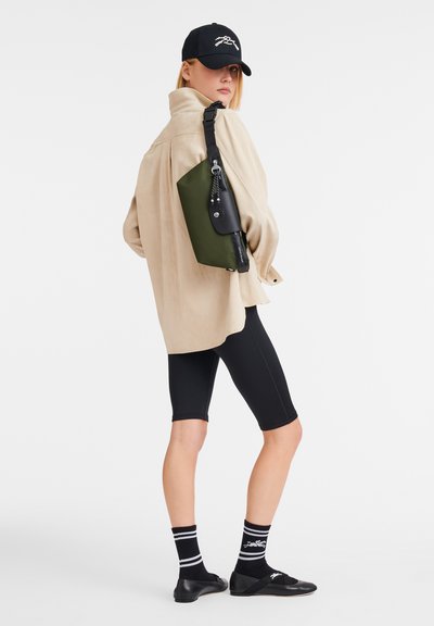 Schwarze Umhängetasche, die an einem grünen Panel befestigt ist, getragen über einem beigen Oversize-Hemd, schwarzen Radshorts und schwarzen Slippern mit gestreiften Socken.