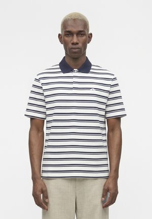 Mand med kort blondt hår iført hvid polo-shirt med tynde marineblå vandrette striber og beige bukser, står mod en hvid baggrund.