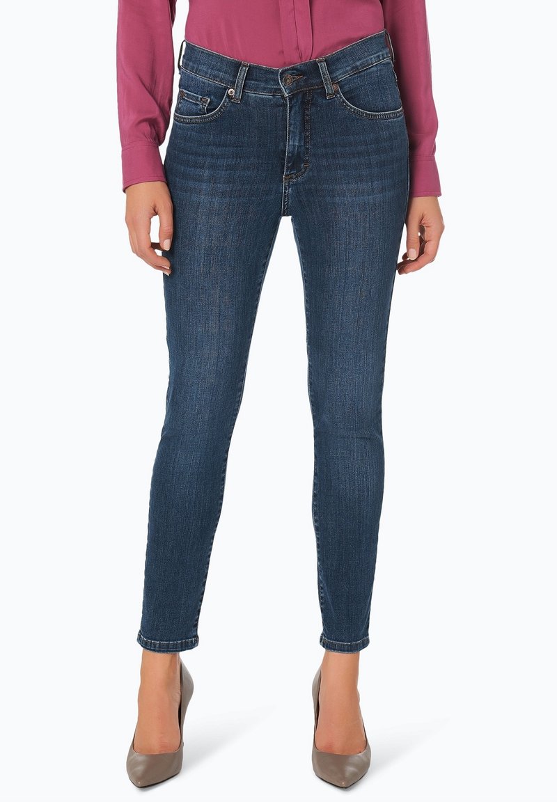 Femme portant un jean skinny bleu foncé, un chemisier rose à manches longues et des chaussures à talons gris, debout devant un fond blanc.
