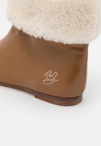 Bottine en cuir marron avec un col en fausse fourrure crème, présentant un logo texturé sur le côté et un talon en bois plat.