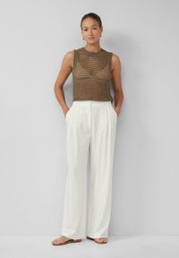Top senza maniche in maglia aperta marrone abbinato a pantaloni bianchi a vita alta. Il modello sta in piedi con le mani nelle tasche, indossando sandali semplici.