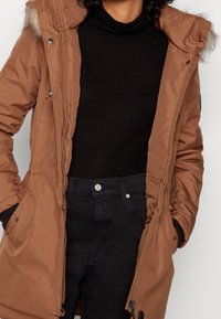 Brun polstret jakke med pelsforet hætte, lynlåslukning og elastisk talje. Båret over en sort rullekrave og sorte jeans.