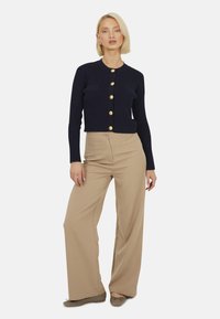 Marineblauwe ribbeltrui met gouden knopen, verkorte lengte, twee voorzakken; gecombineerd met beige wijde broek en platte bruine schoenen.