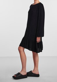 Vestido negro de manga larga con un dobladillo acampanado y volantes, hecho de tejido ligero. Combinado con sandalias de plataforma negras.