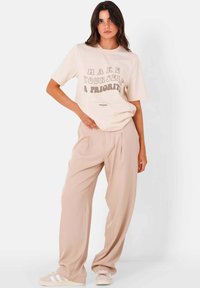 Beige katoenen T-shirt met de tekst "MAAK VAN JOUWZELF EEN PRIORITEIT", gecombineerd met loszittende beige broek en lichtgekleurde sneakers.