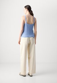 Lys blå strikket tanktop med brede stropper, teksturert design og knappedetalj; kombinert med kremfargede baggy bukser og beige sko.