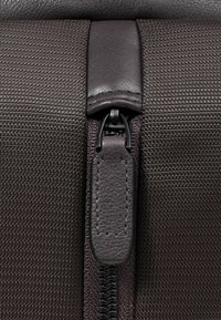 Sezione con cerniera in tessuto marrone scuro texturizzato, dotata di linguetta di pelle e robusta cerniera metallica nera.