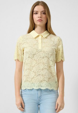 Jeune femme portant un polo à manches courtes en dentelle florale jaune pâle et un jean bleu clair, debout devant un fond uni.