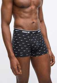 Μυώδης άνδρας που φοράει μαύρα μπόξερ της Nike με λευκά σχέδια Swoosh και λωρίδα μέσης με το σήμα της εταιρείας, στέκεται μπροστά σε απλό φόντο.