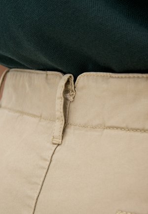 Beige Baumwollhose mit glatter Textur und genähtem Bund, ausgestattet mit einer kleinen Stoffschlaufe zum Aufhängen. Ein dunkelgrüner Pullover ist teilweise sichtbar.