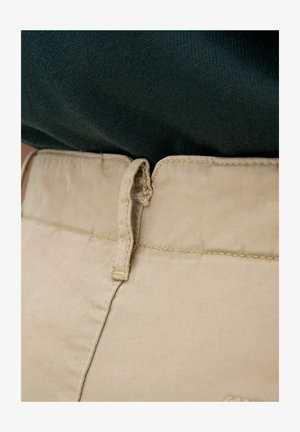 Beige Baumwollhose mit glatter Textur und genähtem Bund, ausgestattet mit einer kleinen Stoffschlaufe zum Aufhängen. Ein dunkelgrüner Pullover ist teilweise sichtbar.