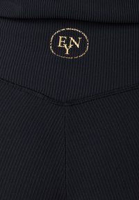 Il tessuto a coste nero è decorato con un'emblema dorata. Il design include una cucitura a zigzag in vita, che aggiunge texture e dimensione.