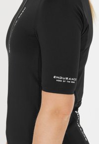 Schwarzes Sportshirt mit kurzen Ärmeln, ausgestattet mit einem Frontreißverschluss, weißem Logotext am Arm und einer taillierten Passform für Sportbekleidung.