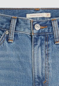 Pantalones cortos anchos de mezclilla azul con costuras naranjas, cierre con botón metálico y una etiqueta que dice '94 Baggy Shorts' talla 27.