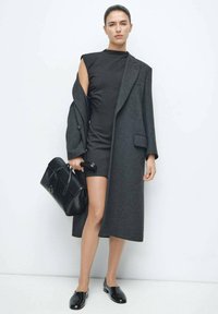 Manteau gris en laine avec revers crantés, porté sur une mini robe noire côtelée. Accessoires : un sac à main noir et des mocassins noirs brillants.