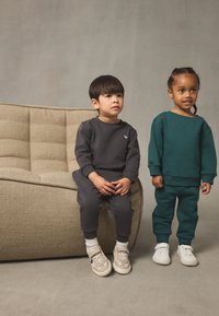 Deux enfants portent des sweats à capuche et des pantalons de survêtement assortis ; l'un est gris foncé et l'autre bleu sarcelle profond. Ils se tiennent à côté d'un canapé beige texturé.