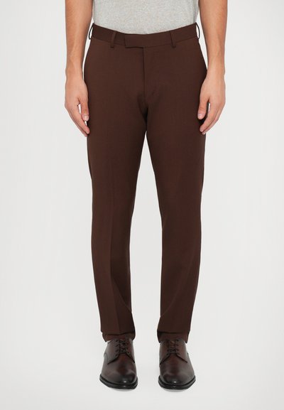 Pantaloni maro personalizați, cu o textură netedă, croială slim și bretele pentru centură, purtați peste pantofi maro din piele. Partea de sus stilată de culoare gri este vizibilă.