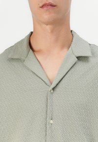 Camisa de manga corta verde claro con tejido texturizado, cuello y tres botones en la parte delantera. Diseño casual y transpirable.