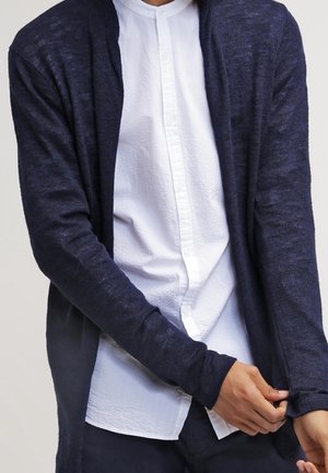 Gilet en tricot marine avec un devant ouvert, des manches longues et des poignets côtelés, superposé à une chemise blanche à boutons texturée.