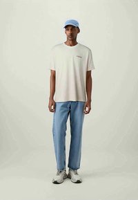 Man wearing light beige oversized t-shirt, light blue jeans, grey sneakers, and light blue cap, standing against plain pale background.