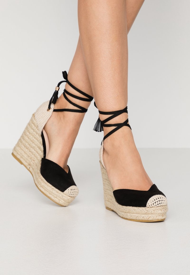 Espadrilles à talons compensés en suede noir avec des accents tissés beiges, des liens à la cheville et un bout rond. Surface texturée avec une semelle en cordes naturelles.