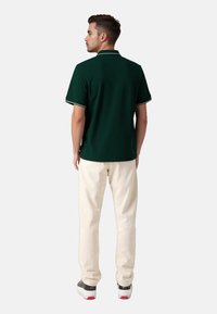 Ciemnozielona koszulka polo z białym kołnierzykiem i mankietami, zestawiona z jasnymi spodniami. Model stoi tyłem do aparatu.