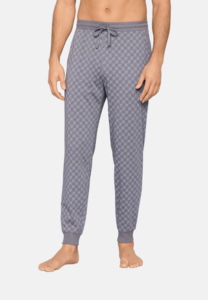 Mandlig model iført grå joggingbukser med et subtilt geometrisk mønster, snøre i taljen og manchetter ved anklerne, stående barfodet på en hvid baggrund.