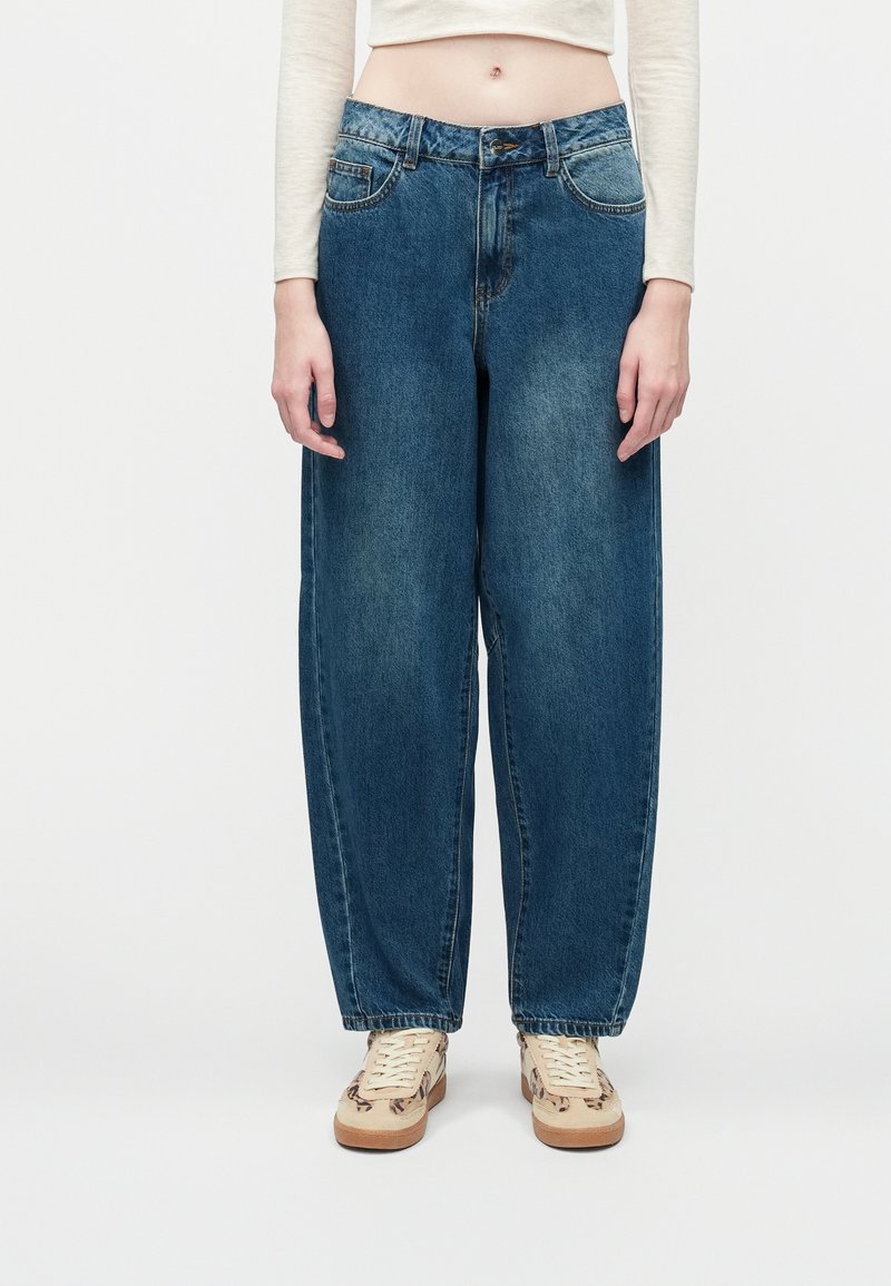Løse blå denimjeans med en lys vask, fem lommer og lukning med knap. Parret med beige sneakers med et subtilt mønster.