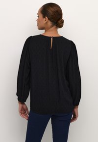 Blouse noire avec un tissu texturé, des manches bouffantes froncées, un décolleté en trou de serrure dans le dos, et une coupe décontractée. Associée à un jean bleu foncé.