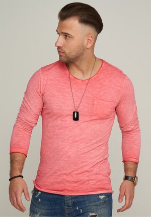 Langärmliges, meliertes korallenfarbenes T-Shirt mit Rundhalsausschnitt und einer Brusttasche, kombiniert mit verwaschenen blauen Jeans und einer schwarzen Anhängerkette.