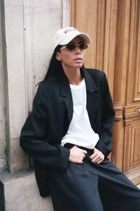 Musta ylipitkä bleiseri, valkoinen t-paita, leveät mustat housut, beige lippalakki, jossa on brodeerattu "WELL", pyöreät aurinkolasit, sileät hiukset, puinen tausta.