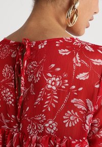 Robe à motif floral rouge avec un tissu texturé, présentant une finition à volants et une fermeture à nouer à l'arrière. Boucle d'oreille en or visible.