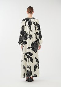 Robe longue et fluide de couleur crème avec des motifs floraux noirs. Elle présente des manches longues et amples ainsi qu'un col resserré. Vue de dos présentée.