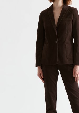 Femme portant un blazer marron foncé cintré et un pantalon assorti, debout devant un fond clair uni, les mains relâchées le long du corps.