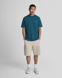 Tirkīza kokvilnas T-krekls ar apaļu apkakli un dzeltenu akcents logotipu, kopā ar bēšām kravas šortām ar sānu kabatām un baltām zeķēm pelēkos apavos.