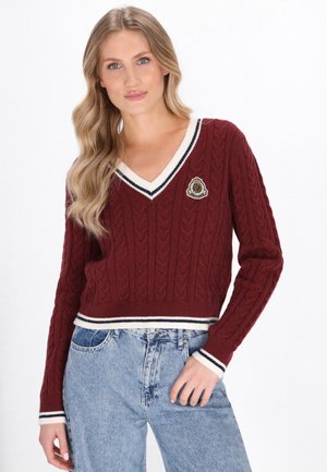 Bordeaux kabelgebreide pullover met V-hals, crèmewitte gestreepte afwerking en geborduurd patch; gecombineerd met hoogopgetrokken lichtblauwe jeans.