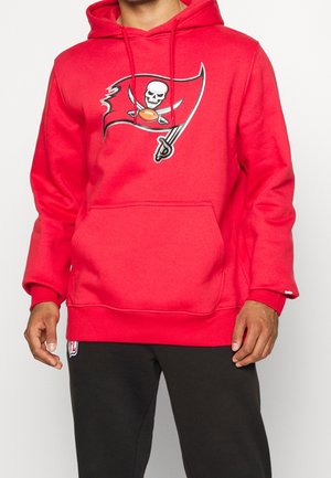 Mężczyzna w czerwonej bluzie z kapturem z logo pirackiej flagi przedstawiającej czaszkę, skrzyżowane miecze i piłkę nożną na piersi, w połączeniu z czarnymi spodniami.