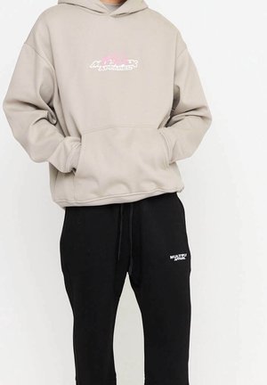 Personne portant un sweat à capuche beige avec un logo rose et blanc et un pantalon de survêtement noir avec un logo blanc, les mains dans la poche du sweat.